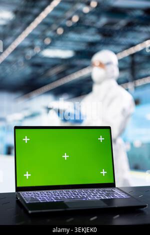 Ordinateur portable à écran vert dans la salle blanche de l'usine PCB utilisé pour surveiller la télémétrie de l'équipement des lignes de production. Gros plan d'une maquette d'ordinateur portable utilisée par un ingénieur dans un équipement de sécurité pour documenter les variations de rendement des micropuces Banque D'Images