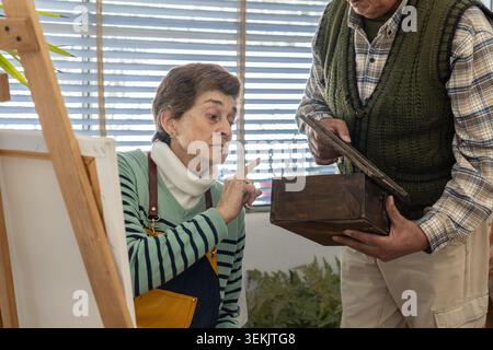 Focused Older Woman pointe vers une boîte en bois sombre tenue par un homme, l'étudiant soigneusement comme référence pour ses œuvres d'art dans un studio lumineux. Banque D'Images