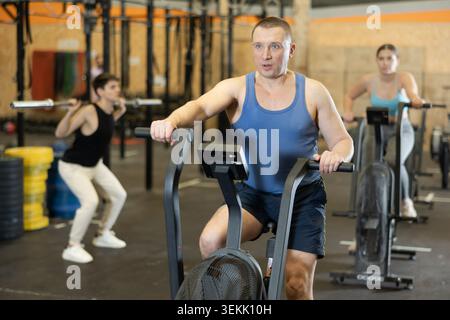 Homme d'âge moyen actif et actif faisant de l'entraînement cardio sur un vélo d'appartement comme un entraînement multidisciplinaire dur dans la salle de gym à l'intérieur Banque D'Images