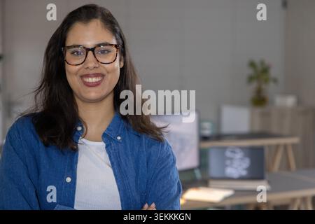 Femme dans les années 20 debout souriante, bras croisés, en chemise en denim au bureau avec ordinateur portable et lunettes Banque D'Images