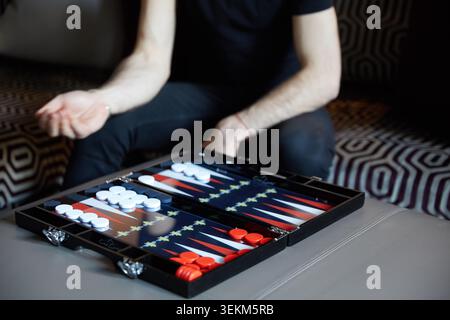 Gros plan d'une planche de backgammon noir de luxe avec des dames rouges et blanches lors d'un jeu d'intérieur sur un pouf en cuir. Banque D'Images