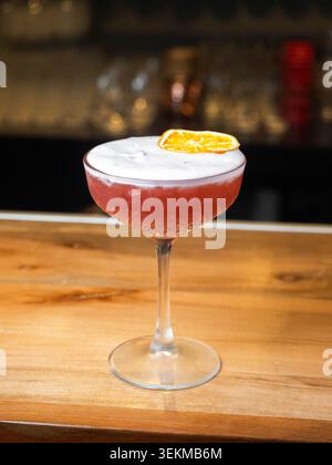 Cocktail rouge avec garniture orange sur un bar en bois. La mousse couronne la boisson, ajoutant un attrait visuel. Un éclairage chaud améliore les tons de couleurs riches. Parfait pour Banque D'Images