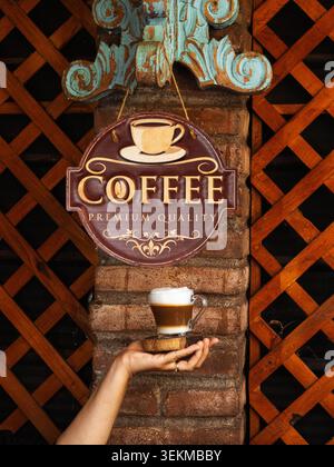 La main tient la tasse de café, chaude et accueillante. Panneau accroche au-dessus, rustique et charmant. Mur de briques et scène de cadre en treillis en bois. La tasse présente un latte crémeux, Banque D'Images