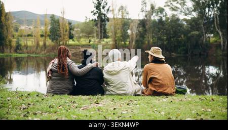Amis, détendez-vous et parlez au parc au bord du lac, point ou embrasser sur le voyage de randonnée, pause et tourisme de la nature. Personnes, groupe et voyage pour vue sur herbe, barrage Banque D'Images