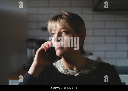 Femme avec la frange regardant loin tout en parlant sur téléphone intelligent à la maison Banque D'Images