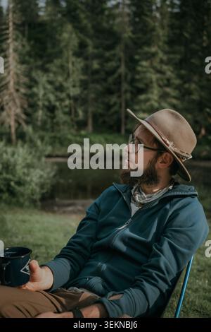 Homme contemplatif tenant une tasse assise sur une chaise de camping tout en regardant loin dans la forêt Banque D'Images