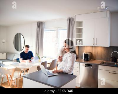 Mère freelance travaillant sur ordinateur portable tout en se tenant debout avec bébé garçon à la maison Banque D'Images