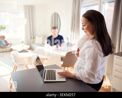 Femme debout avec son fils tout-petit tout en travaillant de la maison sur ordinateur portable Banque D'Images