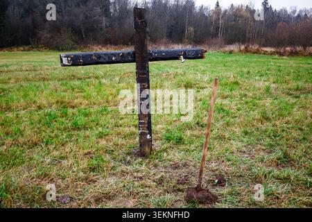 Une croix en bois grossièrement faite et altérée se dresse dans un champ herbeux avec une pelle à côté. Le bois sombre et brûlé suggère un mémorial sombre ou improvisé Banque D'Images