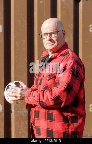 Ouvrier chauve adulte debout avec une balle à la main, expression souriante sur le visage, mur en tôle ondulée en arrière-plan, vêtu de façon décontractée de rouge et de bla Banque D'Images