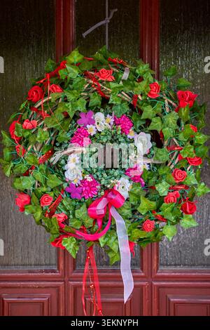 Couronne de porte florale avec des feuilles de lierre vert, des roses rouges, des fleurs roses et blanches, et des rubans colorés suspendus sur une porte d'entrée en bois. Banque D'Images