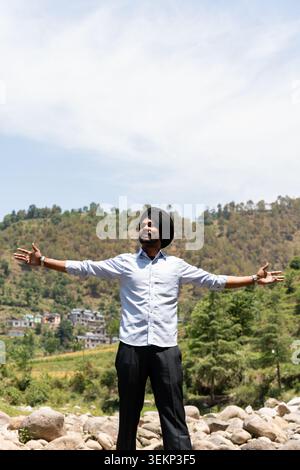 Vue verticale d'un jeune homme sikh punjabi portant un turban noir se tient avec les bras grands ouverts, regardant vers le ciel sur un fond panoramique. Banque D'Images