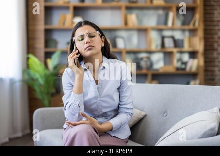 Stressée jeune femme portant des lunettes se sentant fatiguée et frustrée, ayant mal à la tête en étant assise sur un canapé et parlant sur un smartphone pendant un appel téléphonique difficile à domicile à distance Banque D'Images