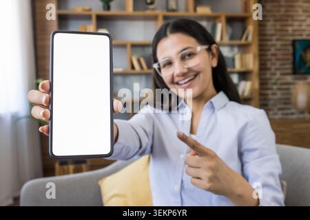 Jeune femme souriante tenant un téléphone portable avec un écran blanc vierge et pointant vers lui, parfait pour présenter de nouvelles applications, promotions ou contenu numérique à la maison Banque D'Images