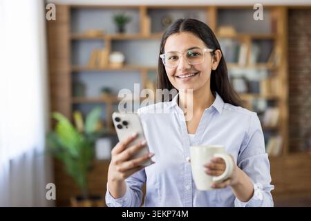 Jeune femme indienne portant des lunettes et une chemise décontractée d'affaires, souriante tout en tenant un smartphone et une tasse à café dans un bureau moderne ou un bureau avec des étagères en arrière-plan Banque D'Images
