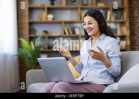 Jeune femme portant des lunettes en riant et en faisant des gestes lors d'une visioconférence en ligne, assise confortablement sur un canapé à la maison tout en communiquant avec des collègues Banque D'Images