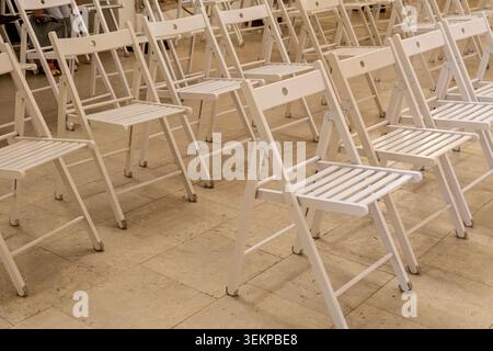 Une vue en perspective à grand angle de plusieurs rangées de chaises pliantes blanches vides en bois soigneusement disposées sur un sol en pierre pour une cérémonie publique en plein air ou Banque D'Images