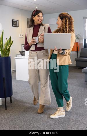 Deux femmes professionnelles marchant et parlant à travers le bureau moderne, portant ordinateur portable et tasse à café Banque D'Images