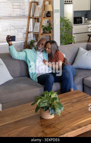 Couple senior diversifié assis à proximité sur le canapé sectionnel gris, prenant selfie avec smartphone Banque D'Images