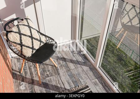 scène de balcon confortable avec une chaise noire, un oreiller à motifs et des carreaux de sol de style bois, baigné de lumière du soleil Banque D'Images