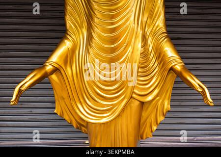 Gros plan détaillé d'une statue dorée de Bouddha, mettant en évidence les mains gracieusement étendues et les plis complexes de la robe monastique, dressée contre Banque D'Images