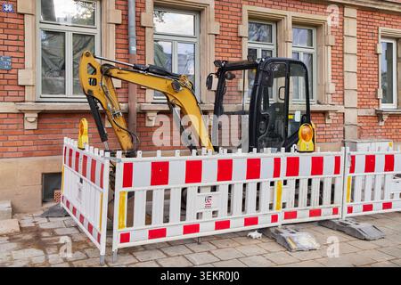 Une petite pelle jaune stationnée derrière des barrières de sécurité rouges et blanches sur un chantier de construction de rue en ville. Banque D'Images