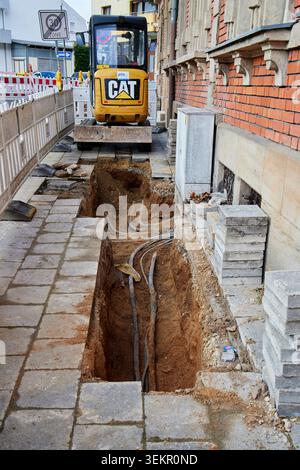 Une petite pelle Cat jaune stationnée derrière une tranchée profonde avec des tuyaux exposés sur un chantier de construction de rue urbaine. Banque D'Images