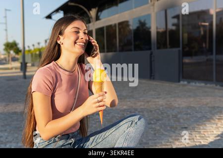 Femme profitant d'une conversation téléphonique tout en tenant un cône de crème glacée à la mangue dans son autre main. Banque D'Images