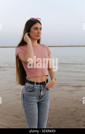 Une femme aux cheveux longs se tient sur le rivage se relaxant et profitant de ses vacances. Elle porte des lunettes de soleil et a la main sur la hanche Banque D'Images