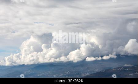 Massifs, spectaculaires nuages de cumulus blancs et moelleux se rassemblant bas au-dessus d'une vallée de montagne bleu brumeux avec de petits villages à flanc de colline dispersés en dessous. Banque D'Images