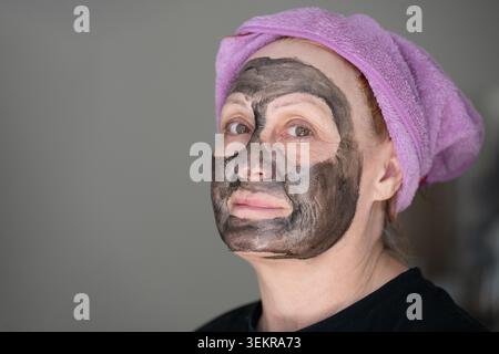 Portrait d'une femme d'âge moyen avec un masque de spa noir purifiant sur son visage regardant la caméra. Femme âgée prenant soin de sa peau appliquant cosmétique Banque D'Images