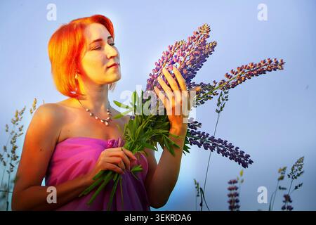 Femme en tenue violette appréciant les lupins violets dans un champ de floraison pendant l'heure dorée en été Banque D'Images
