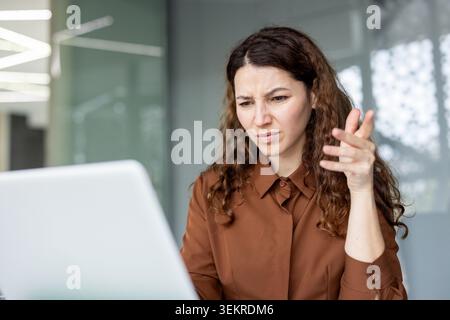 Jeune femme assise à un bureau dans un bureau moderne, paraissant perplexe et faisant des gestes vers son ordinateur portable pendant une tâche de travail en ligne Banque D'Images