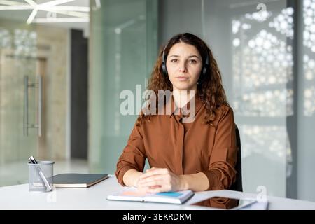 Une femme aux cheveux bouclés portant des écouteurs est assise à un bureau dans un bureau moderne lumineux, face à la caméra avec une expression calme et attentive et des cahiers dedans Banque D'Images