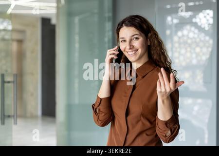 Jeune femme souriante debout à l'intérieur dans un bureau moderne et parlant sur un smartphone tout en faisant des gestes avec son autre main, avec un espace de travail vitré lumineux Banque D'Images