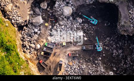 Vue aérienne d'une carrière de pierre avec des excavatrices et des camions à benne basculante travaillant parmi de grands rochers et des ouvriers sur le sol rocheux en Indonésie. Banque D'Images