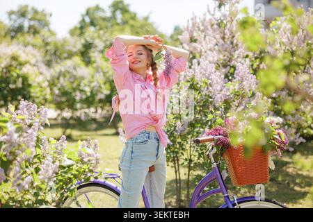 Une jeune femme joyeuse avec des fleurs dans les cheveux pose à côté de son vélo dans un jardin animé, incarnant l'essence du cyclisme et des loisirs actifs Banque D'Images