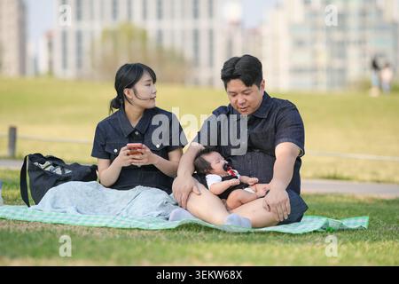 La famille profite d'un moment de parc calme ensemble. Mère tient le téléphone, père berce bébé. L'herbe douce et les bâtiments éloignés encadrent la scène. La sensibilité rayonne f Banque D'Images