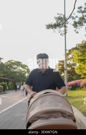 L'homme pousse la poussette le long du chemin pavé du parc. Arbres et fleurs bordent la passerelle ensoleillée. Une lumière douce illumine son expression focalisée. Un moment paisible Banque D'Images