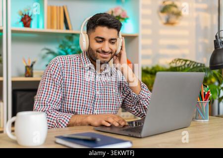 L'homme indien sur la table à la maison travaille sur ordinateur portable dans des écouteurs sans fil tapant du texte et écoutant de la musique avec une concentration calme à l'intérieur. Arabian Guy sur la table de la maison apprécie le travail freelance détendu et la routine de bureau à domicile Banque D'Images