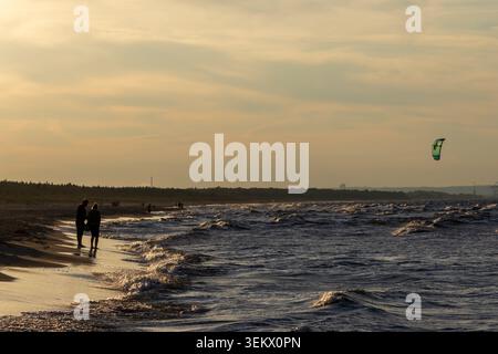 Sobiszewska Island, POLOGNE-SEPTEMBRE 02, 2024 : couple marchant le long d'une plage de sable au coucher du soleil, profitant de la mer Baltique ondulée tandis qu'un kite surfeur est coloré Banque D'Images