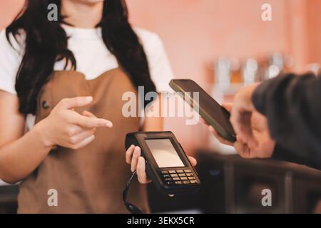 Barista adulte senior asiatique recevant un paiement mobile de la part d'un client utilisant un smartphone dans un café démontrant une transaction sans espèces moderne dans Cozy lo Banque D'Images