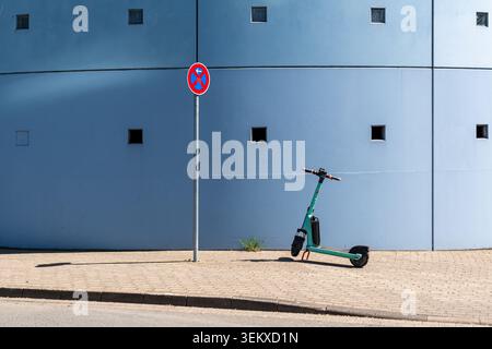 Un seul scooter électrique vert partagé (e-scooter) est garé sur un trottoir de la ville à côté d'un panneau de non stationnement contre un mur urbain bleu minimal. Banque D'Images