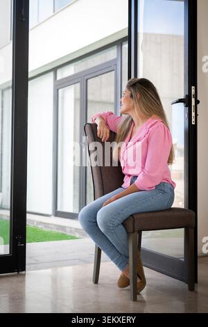 Femme détendue assise sur une chaise près d'une grande fenêtre à la maison, regardant dehors avec une expression réfléchie. Concept de style de vie lié à la pleine conscience, Banque D'Images