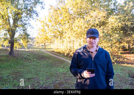 Homme adulte en veste bleue et casquette tenant un smartphone à l'extérieur sur un chemin avec des arbres Banque D'Images
