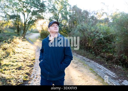 Homme dans une veste bleue et casquette marchant un chemin extérieur, levant les yeux avec une expression contemplative Banque D'Images