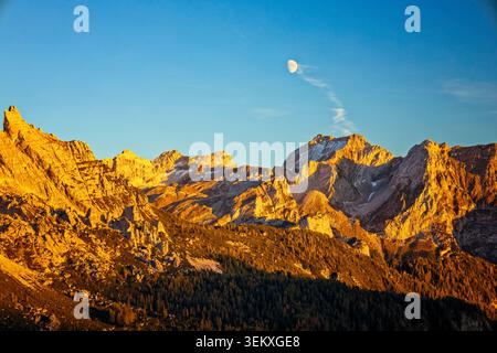 La Valle Agordina, Italie - 15.10.2021 : majestueux sommets rocheux des Dolomites baignés à alpenglow au-dessus de forêts de pins sombres sous une lune pâle Banque D'Images