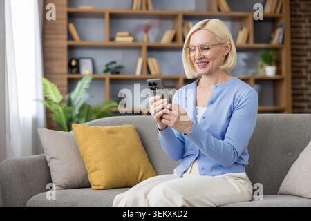 Femme blonde d'âge moyen dans des lunettes assise sur un canapé confortable du salon, souriante et détendue tout en naviguant et en envoyant des messages sur son smartphone, en profitant de la connexion numérique et des loisirs Banque D'Images