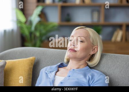 Femme senior se détache à la maison sur le canapé, fermant les yeux dans la contemplation sereine, pratiquant la respiration consciente pour la paix intérieure et le bien-être mental dans un cadre confortable de salon Banque D'Images