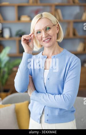 Femme mature joyeuse souriante et ajustant ses lunettes, debout avec confiance dans un bureau à domicile moderne ou un cadre de bibliothèque, symbolisant l'intelligence, l'expérience et le professionnalisme accessible Banque D'Images